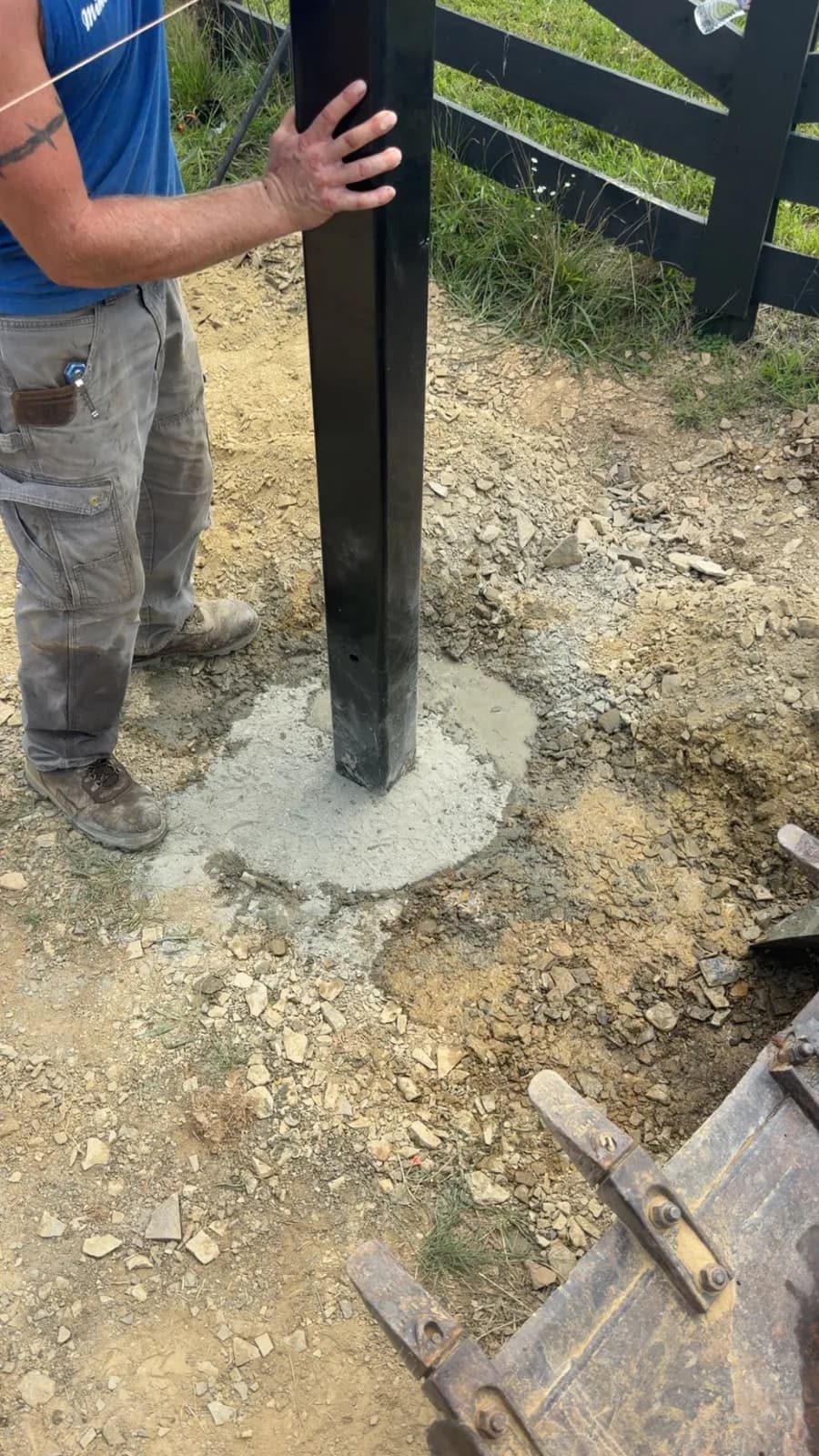 Steel gate post set plumb in fresh concrete on a black board-fence line