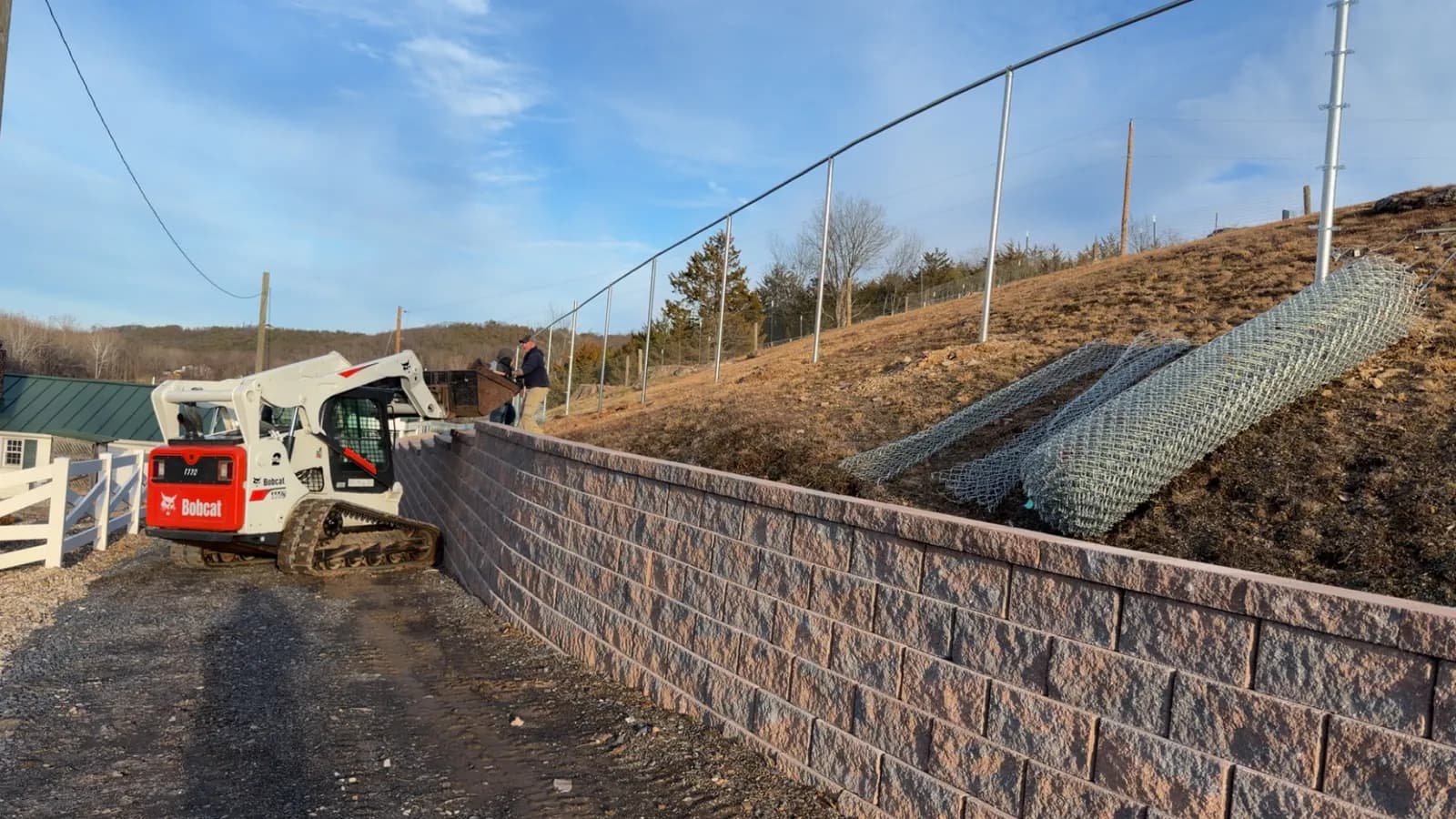 Compact track loader staged on a retaining wall during a chain-link install